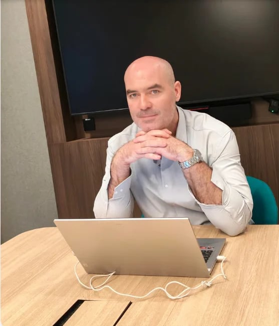 Professional man in light blue shirt sitting at desk with laptop, resting chin on hands in thoughtful pose